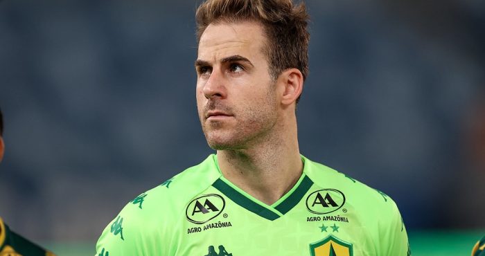 “Jogador do Cuiabá Esporte Clube durante atividade de treinamento, segurando a bola com firmeza, com uniforme azul e luvas de goleiro, em campo de treino ao ar livre.”