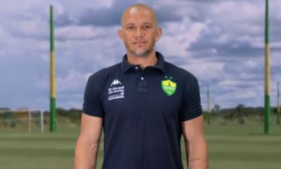Imagem do novo técnico do Cuiabá Sub-20, Nei Cardoso, vestindo a camisa do clube. Ele está sorrindo e posando para a foto, com o campo de treinamento ao fundo.