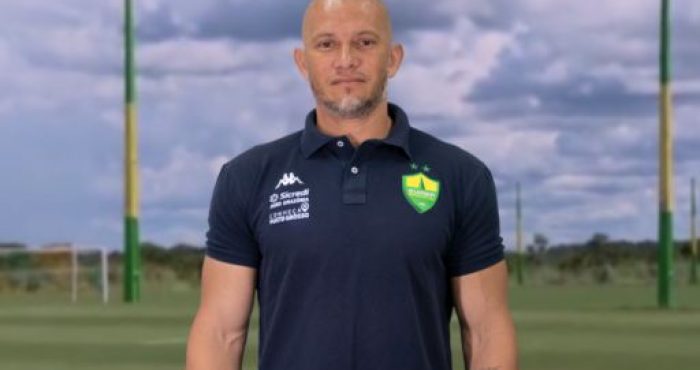Imagem do novo técnico do Cuiabá Sub-20, Nei Cardoso, vestindo a camisa do clube. Ele está sorrindo e posando para a foto, com o campo de treinamento ao fundo.