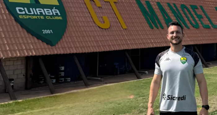 Lucas Barezani posa sorrindo em frente ao prédio do CT Manoel Dresch, centro de treinamento do Cuiabá Esporte Clube, vestindo camisa oficial de treino do clube.