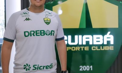 Silvio Eduardo, novo reforço do departamento de futebol do Cuiabá Esporte Clube, posa em frente ao escudo do clube vestindo a camisa oficial do Dourado.