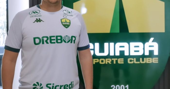 Silvio Eduardo, novo reforço do departamento de futebol do Cuiabá Esporte Clube, posa em frente ao escudo do clube vestindo a camisa oficial do Dourado.