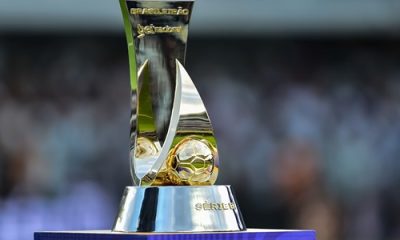 Troféu da Série B do Campeonato Brasileiro em destaque sobre um pedestal, com fundo desfocado de um estádio e torcida. O troféu possui design moderno com detalhes dourados e uma bola em relevo na base.