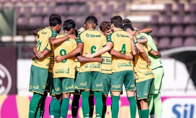 Jogadores do time sub-20 do Cuiabá reunidos em um círculo (perfilado de costas) no centro do gramado antes do início da partida pela Copa São Paulo de Futebol Júnior. Eles vestem o uniforme reserva: camisa amarela com detalhes verdes, calções verdes e meiões verdes. É possível ver o patrocinador "Sicredi" e a frase "Conheça Mato Grosso" nas costas das camisas.
