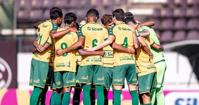 Jogadores do time sub-20 do Cuiabá reunidos em um círculo (perfilado de costas) no centro do gramado antes do início da partida pela Copa São Paulo de Futebol Júnior. Eles vestem o uniforme reserva: camisa amarela com detalhes verdes, calções verdes e meiões verdes. É possível ver o patrocinador "Sicredi" e a frase "Conheça Mato Grosso" nas costas das camisas.