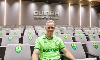 Fotografia do goleiro João Carlos sentado em uma das poltronas de uma sala de imprensa. Ele veste o uniforme verde-limão do Cuiabá e olha para a câmera com um sorriso amigável. As poltronas ao redor são cinzas com o brasão do clube. No topo da parede cinza ao fundo, destaca-se o nome do time em relevo sob iluminação focada.