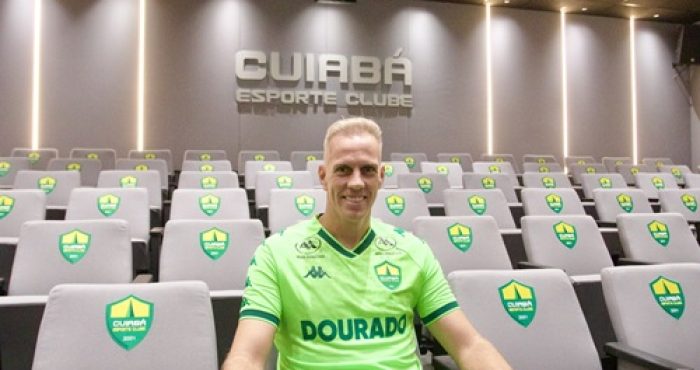 Fotografia do goleiro João Carlos sentado em uma das poltronas de uma sala de imprensa. Ele veste o uniforme verde-limão do Cuiabá e olha para a câmera com um sorriso amigável. As poltronas ao redor são cinzas com o brasão do clube. No topo da parede cinza ao fundo, destaca-se o nome do time em relevo sob iluminação focada.