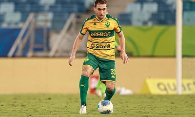 "Jogador do Cuiabá EC em ação durante uma partida, vestindo o uniforme verde e amarelo do clube, conduzindo a bola no gramado da Arena Pantanal."