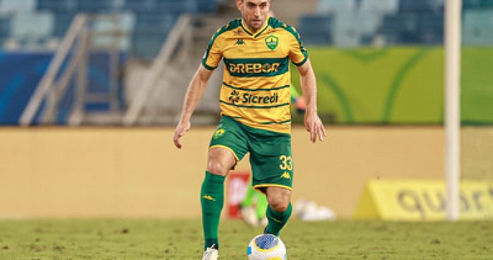 "Jogador do Cuiabá EC em ação durante uma partida, vestindo o uniforme verde e amarelo do clube, conduzindo a bola no gramado da Arena Pantanal."