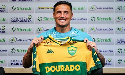 O atacante Edu é apresentado oficialmente como reforço do Cuiabá Esporte Clube. Na imagem, ele sorri enquanto segura a camisa amarela e verde do Dourado durante a coletiva de imprensa, com um painel de patrocinadores ao fundo.