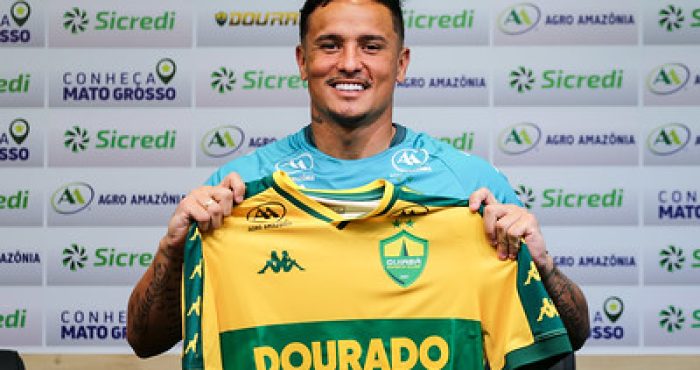 O atacante Edu é apresentado oficialmente como reforço do Cuiabá Esporte Clube. Na imagem, ele sorri enquanto segura a camisa amarela e verde do Dourado durante a coletiva de imprensa, com um painel de patrocinadores ao fundo.