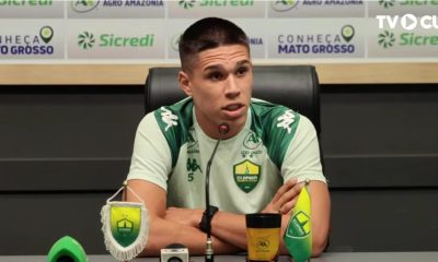 Calebe está sentado à frente de um painel de patrocinadores (backdrops), onde é possível ver marcas como Sicredi, Agro Amazônia e o selo "Conheça Mato Grosso". À sua frente, há microfones e um copo personalizado do clube. No canto superior direito, há a marca d'água da TV Cuiabá, o canal oficial de comunicação do time.