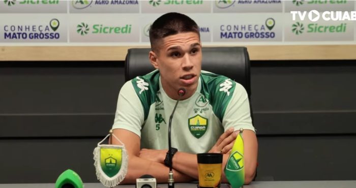Calebe está sentado à frente de um painel de patrocinadores (backdrops), onde é possível ver marcas como Sicredi, Agro Amazônia e o selo "Conheça Mato Grosso". À sua frente, há microfones e um copo personalizado do clube. No canto superior direito, há a marca d'água da TV Cuiabá, o canal oficial de comunicação do time.