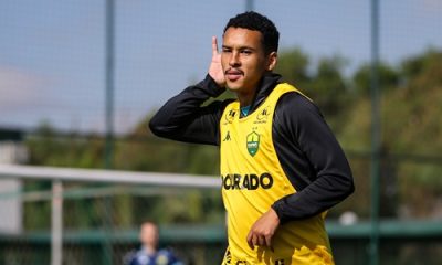 Atleta do Cuiabá com colete de treino amarelo e agasalho preto, comemorando com gesto de mão no ouvido durante atividade no CT. Ao fundo, vê-se o gol e parte da comissão técnica.