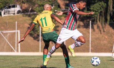 "Jogadores do Cuiabá e do Fluminense disputam a bola durante partida de futebol em campo aberto, com gramado verde e gol ao fundo."