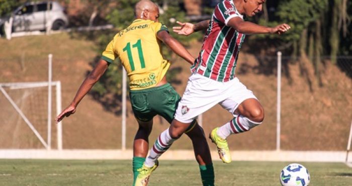 "Jogadores do Cuiabá e do Fluminense disputam a bola durante partida de futebol em campo aberto, com gramado verde e gol ao fundo."