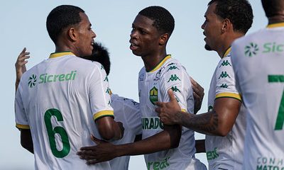Jogadores do Cuiabá Sub-20 comemoram um gol, abraçados e demonstrando emoção. Eles vestem uniformes brancos com detalhes verdes e amarelos, e a logo do Sicredi aparece nas camisas.
