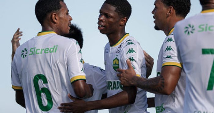 Jogadores do Cuiabá Sub-20 comemoram um gol, abraçados e demonstrando emoção. Eles vestem uniformes brancos com detalhes verdes e amarelos, e a logo do Sicredi aparece nas camisas.