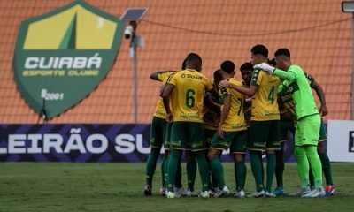 Fotografia em plano médio do grupo de jogadores do Cuiabá-sub20 reunidos em um círculo no gramado durante uma partida. Eles estão de costas para a câmera, vestindo uniformes amarelo e calções verde-escuros. Ao fundo, vê-se o escudo do cuiabá no CT Manoel Dresch.