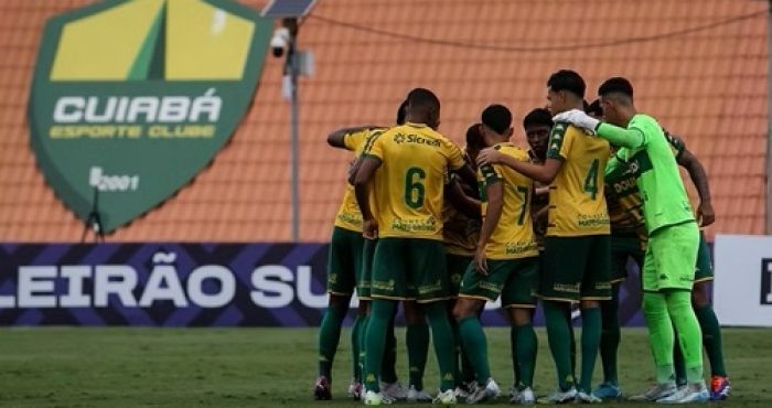 Fotografia em plano médio do grupo de jogadores do Cuiabá-sub20 reunidos em um círculo no gramado durante uma partida. Eles estão de costas para a câmera, vestindo uniformes amarelo e calções verde-escuros. Ao fundo, vê-se o escudo do cuiabá no CT Manoel Dresch.