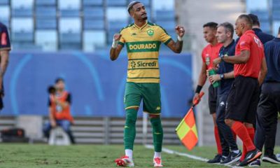 "Derik Lacerda, atacante do Cuiabá, comemora um gol com emoção. Vestindo o uniforme verde e amarelo do Dourado, ele ergue os braços e sorri, enquanto a torcida vibra ao fundo na Arena Pantanal."