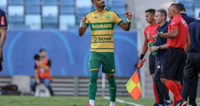 "Derik Lacerda, atacante do Cuiabá, comemora um gol com emoção. Vestindo o uniforme verde e amarelo do Dourado, ele ergue os braços e sorri, enquanto a torcida vibra ao fundo na Arena Pantanal."