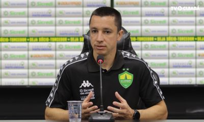 Fotografia de Eduardo Barros, integrante da comissão técnica do Cuiabá Esporte Clube, durante uma entrevista. Ele veste uma camisa polo preta do time e está à frente de um painel com logotipos de patrocinadores como Sicredi e Agro Amazônia.