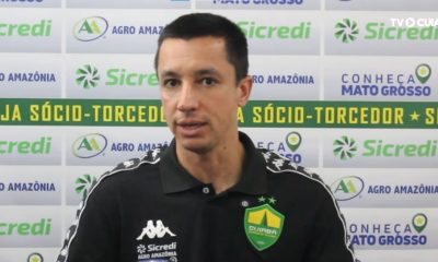 Fotografia de Eduardo Barros, integrante da comissão técnica do Cuiabá Esporte Clube, durante uma entrevista. Ele veste uma camisa polo preta do time e está à frente de um painel com logotipos de patrocinadores como Sicredi e Agro Amazônia.