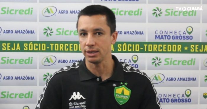 Fotografia de Eduardo Barros, integrante da comissão técnica do Cuiabá Esporte Clube, durante uma entrevista. Ele veste uma camisa polo preta do time e está à frente de um painel com logotipos de patrocinadores como Sicredi e Agro Amazônia.