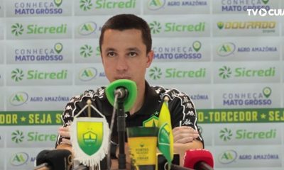 Fotografia de Eduardo Barros, integrante da comissão técnica do Cuiabá Esporte Clube, durante uma entrevista. Ele veste uma camisa polo preta do time e está à frente de um painel com logotipos de patrocinadores como Sicredi e Agro Amazônia.