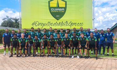 Foto em grupo dos jogadores e comissão técnica do Cuiabá Esporte Clube posando em frente a um grande painel verde e amarelo. O painel exibe o escudo do time e a hashtag "#OrgulhodeMatoGrosso". Os atletas estão organizados em duas fileiras: a da frente agachada e a de trás em pé, todos vestindo o uniforme de treino azul escuro e verde. O ambiente é externo, sob um céu levemente nublado, com árvores ao fundo e um piso de bloquetes de concreto à frente.