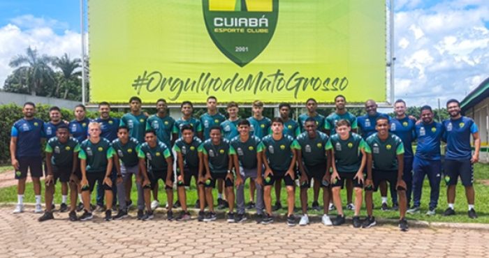 Foto em grupo dos jogadores e comissão técnica do Cuiabá Esporte Clube posando em frente a um grande painel verde e amarelo. O painel exibe o escudo do time e a hashtag "#OrgulhodeMatoGrosso". Os atletas estão organizados em duas fileiras: a da frente agachada e a de trás em pé, todos vestindo o uniforme de treino azul escuro e verde. O ambiente é externo, sob um céu levemente nublado, com árvores ao fundo e um piso de bloquetes de concreto à frente.