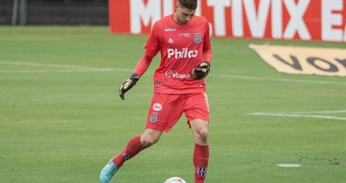 "Goleiro Guilherme Nogueira, do Capivariano, durante partida, vestindo uniforme vermelho e dominando a bola com os pés em campo de futebol."