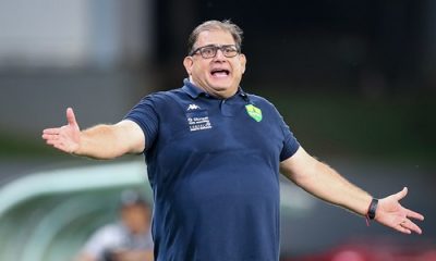 Técnico Guto Ferreira, do Cuiabá, gesticula com os braços abertos durante partida na Arena Pantanal, usando camisa polo azul escura com o escudo do clube no peito e expressão de orientação ou protesto no rosto.