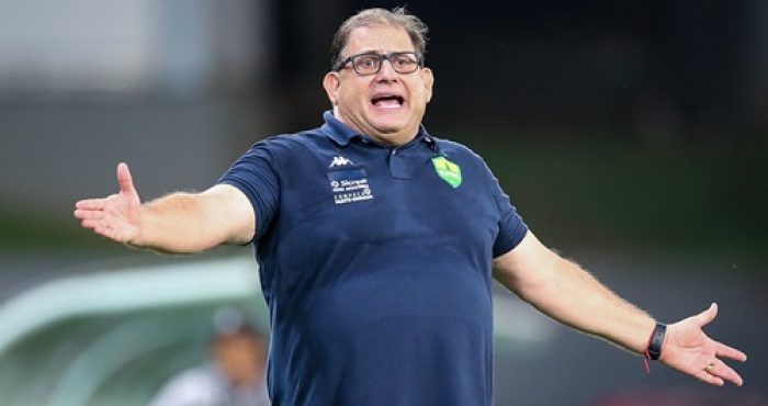 Técnico Guto Ferreira, do Cuiabá, gesticula com os braços abertos durante partida na Arena Pantanal, usando camisa polo azul escura com o escudo do clube no peito e expressão de orientação ou protesto no rosto.