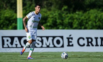 Jogador do Cuiabá Sub-20 conduz a bola durante partida no CT, com faixa "Racismo é Crime" ao fundo.