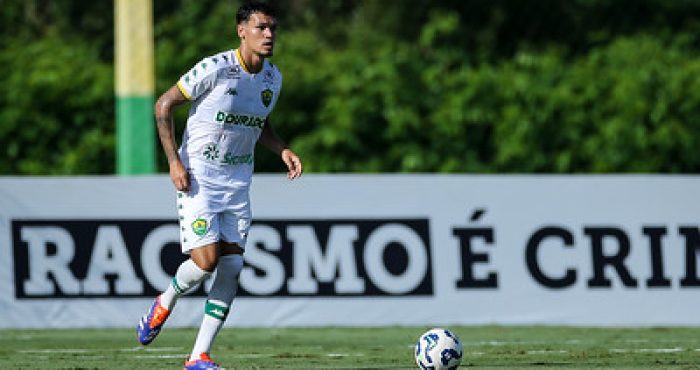 Jogador do Cuiabá Sub-20 conduz a bola durante partida no CT, com faixa "Racismo é Crime" ao fundo.