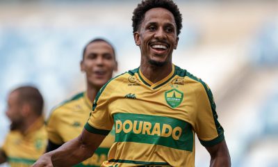 Lucas Mineiro, jogador do Cuiabá, comemora o primeiro gol da partida com os braços erguidos e expressão de felicidade. Ele veste o uniforme auriverde do Dourado, enquanto torcedores vibram ao fundo no estádio.