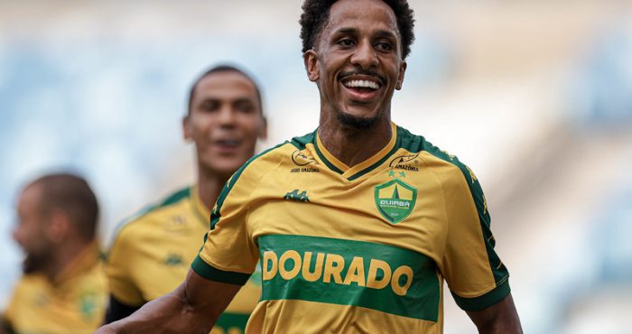 Lucas Mineiro, jogador do Cuiabá, comemora o primeiro gol da partida com os braços erguidos e expressão de felicidade. Ele veste o uniforme auriverde do Dourado, enquanto torcedores vibram ao fundo no estádio.