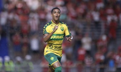 Max comemora seu gol pelo Cuiabá com expressão de alegria, usando o uniforme amarelo e verde, diante da torcida adversária.