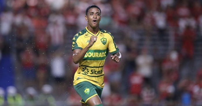 Max comemora seu gol pelo Cuiabá com expressão de alegria, usando o uniforme amarelo e verde, diante da torcida adversária.