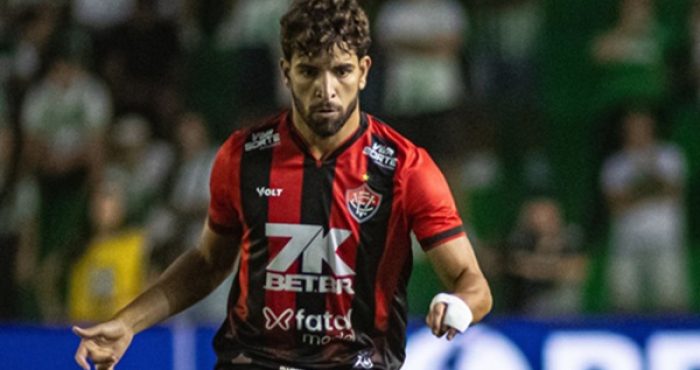 Pepê, volante de futebol, em ação com a camisa do Vitória durante partida, correndo em campo e observando o jogo.