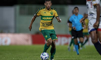 Ruan Oliveira conduz a bola durante partida do Cuiabá, vestindo o uniforme amarelo e verde, com foco e determinação em campo.
