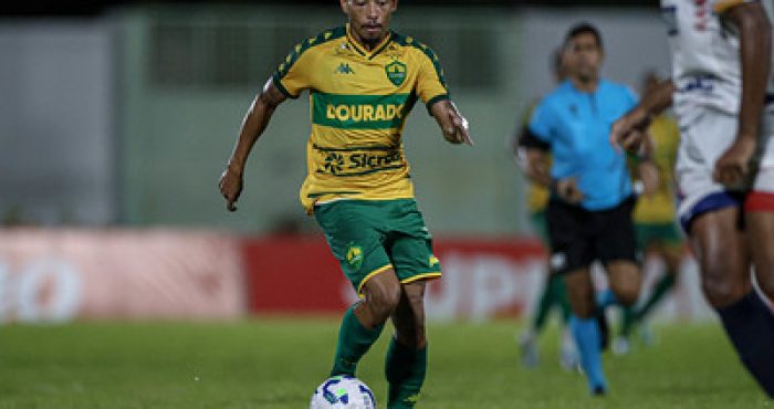 Ruan Oliveira conduz a bola durante partida do Cuiabá, vestindo o uniforme amarelo e verde, com foco e determinação em campo.