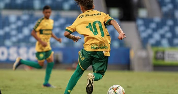 Jogadores do Cuiabá Sub-13 se preparando para a disputa da Liga de Desenvolvimento, com a tabela da competição à vista, simbolizando a entrada do time na elite do futebol infantil.