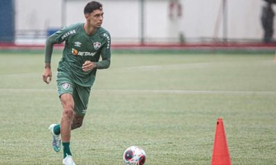 Fotografia de ação do jogador Vitor Mendes durante um treinamento em campo gramado sob chuva leve. Ele está em movimento de corrida, conduzindo uma bola de futebol oficial. Veste um conjunto de treino na cor verde escuro com detalhes brancos e o escudo de um clube anterior. Ao lado dele, vê-se um cone de sinalização laranja no gramado.