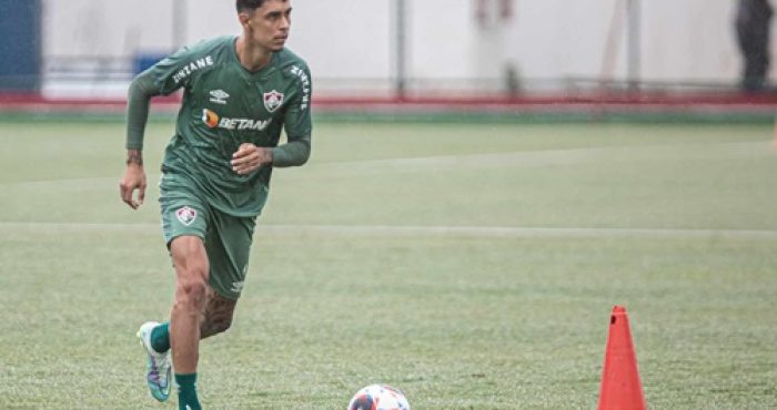 Fotografia de ação do jogador Vitor Mendes durante um treinamento em campo gramado sob chuva leve. Ele está em movimento de corrida, conduzindo uma bola de futebol oficial. Veste um conjunto de treino na cor verde escuro com detalhes brancos e o escudo de um clube anterior. Ao lado dele, vê-se um cone de sinalização laranja no gramado.