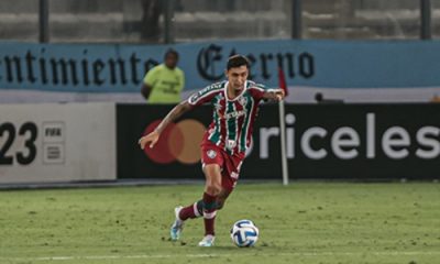 O zagueiro Vitor Mendes, vestindo o uniforme tricolor do Fluminense, conduz a bola com o pé direito durante uma partida de futebol no estádio.
