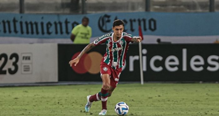O zagueiro Vitor Mendes, vestindo o uniforme tricolor do Fluminense, conduz a bola com o pé direito durante uma partida de futebol no estádio.