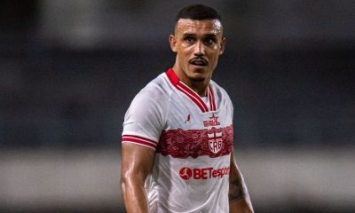 "Fotografia colorida do jogador William Pottker vestindo o uniforme branco do CRB. Ele é um homem pardo, com cabelo curto cortado em degradê e um bigode fino. O atleta aparece suado, com expressão focada, olhando para o lado durante uma partida noturna. A camisa possui detalhes em vermelho na gola e nas mangas, com o escudo do clube e o patrocínio 'BETesporte' em destaque."